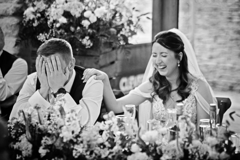 Candid photo of Groom holding his head in his hands with his bride laughing during wedding speeches at Cotswold Venue.