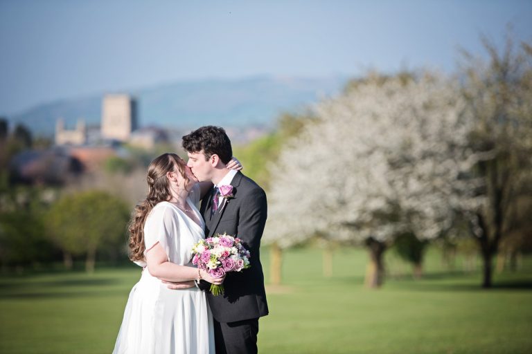 Bride and groom kiss in the grounds at Tewkesbury Park with Tewkesbury Abbey behind them.