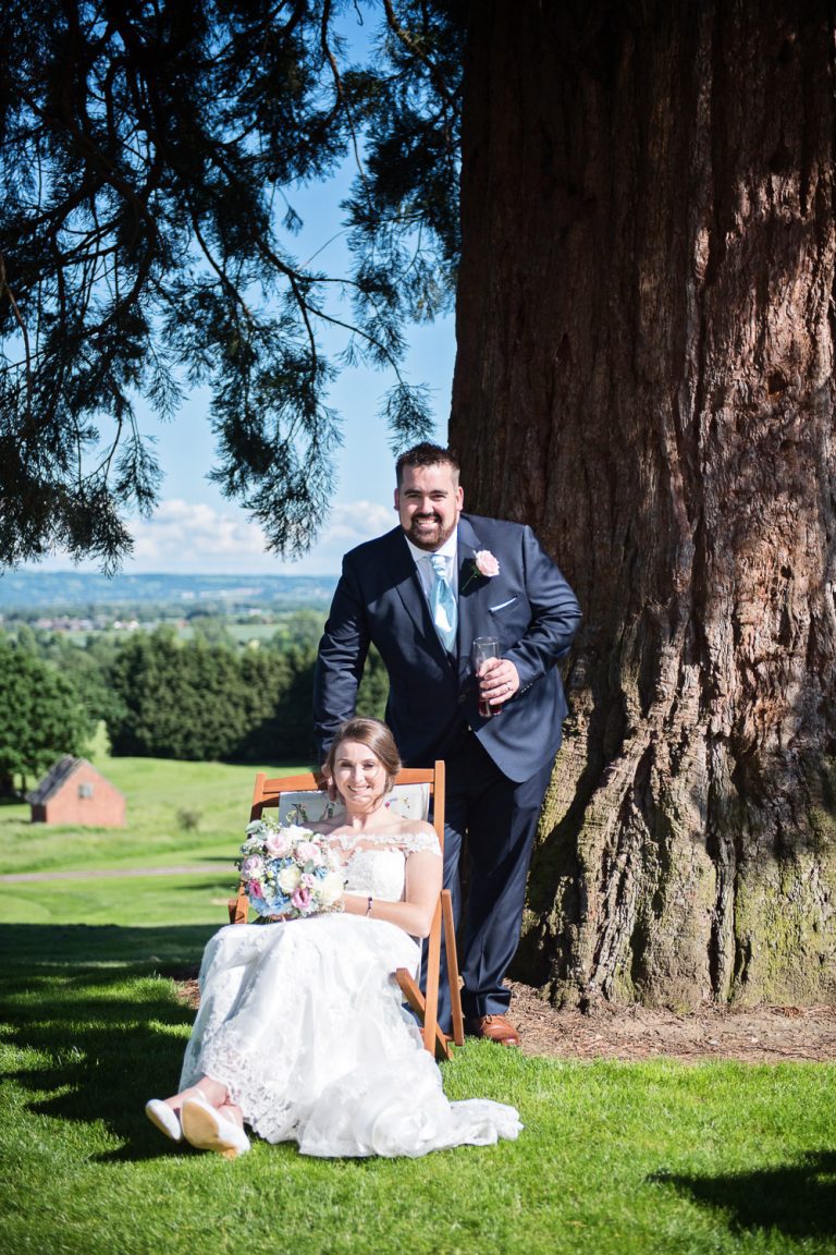 Relaxed image of bride sitting on a deckchair, groom standing behind at Tewkesbury Park