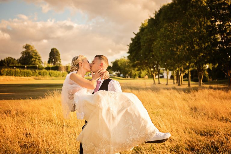 Groom carrying bride in his arms at Tewkesbury Park during sunset.