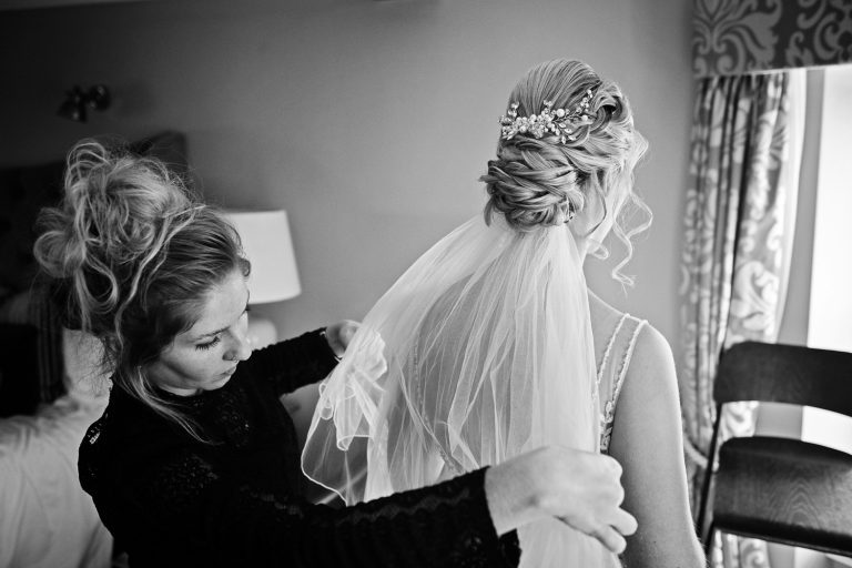 B&W documentary wedding photo of bride having her wedding veil fitted in the bridal suite at Tewkesbury Park.