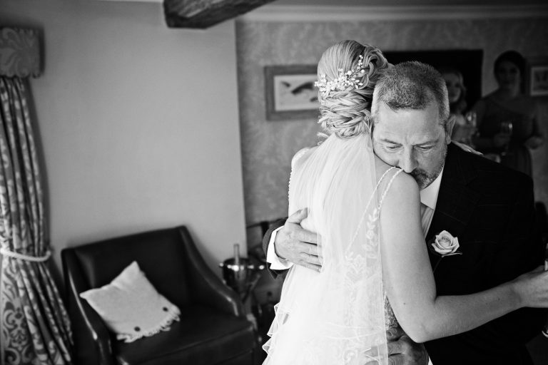 B&W documentary wedding photo of bride being hugged by her father.